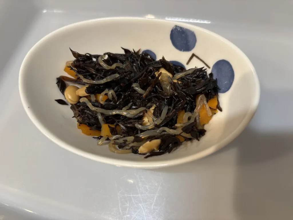 白い楕円の器に盛り付けたひじきの煮物。ひじき・にんじん・糸こんにゃく・大豆が入っている