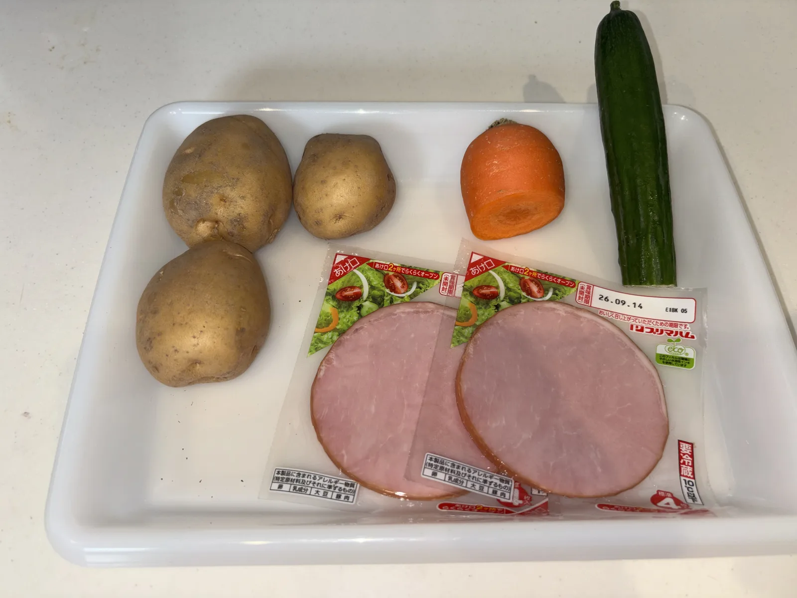 白いトレーに並べた材料一式。じゃがいも3個、ロースハム、にんじん、きゅうり